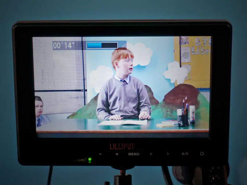A school pupil sits at a desk with a mountain landscape behind him, viewed through a camera display screen.