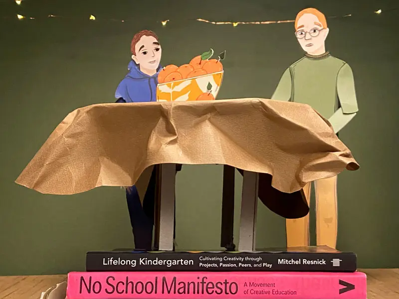 a behind the scenes view of the set for the clan childlaw animation. two characters sit at a table , we see a stack of books stabilising the table.