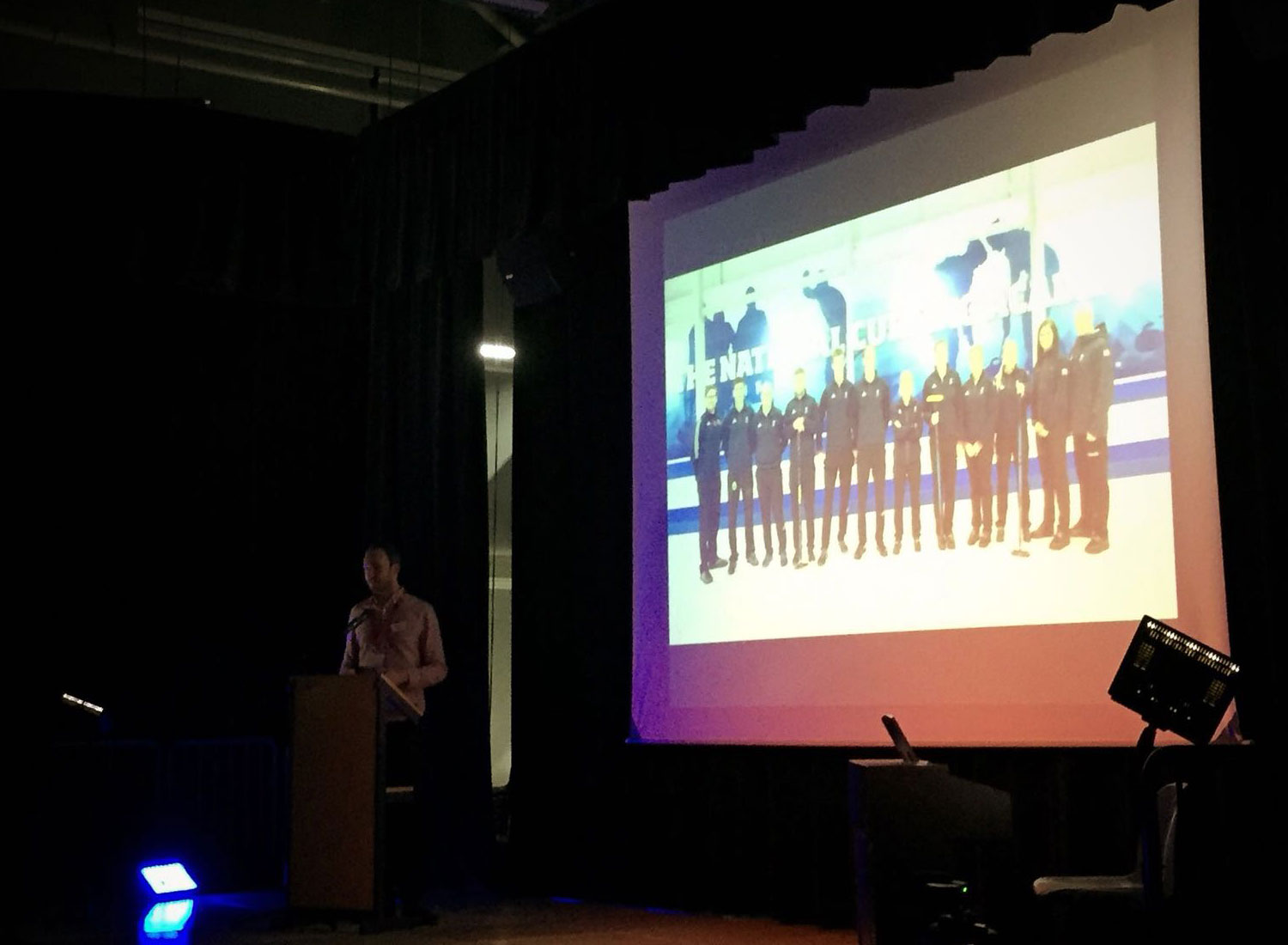 a man talking on stage in front of a projection.